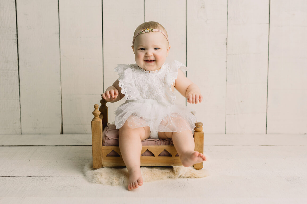 Smiling baby girl in white lace dress on wooden bed prop, Boise baby photographer