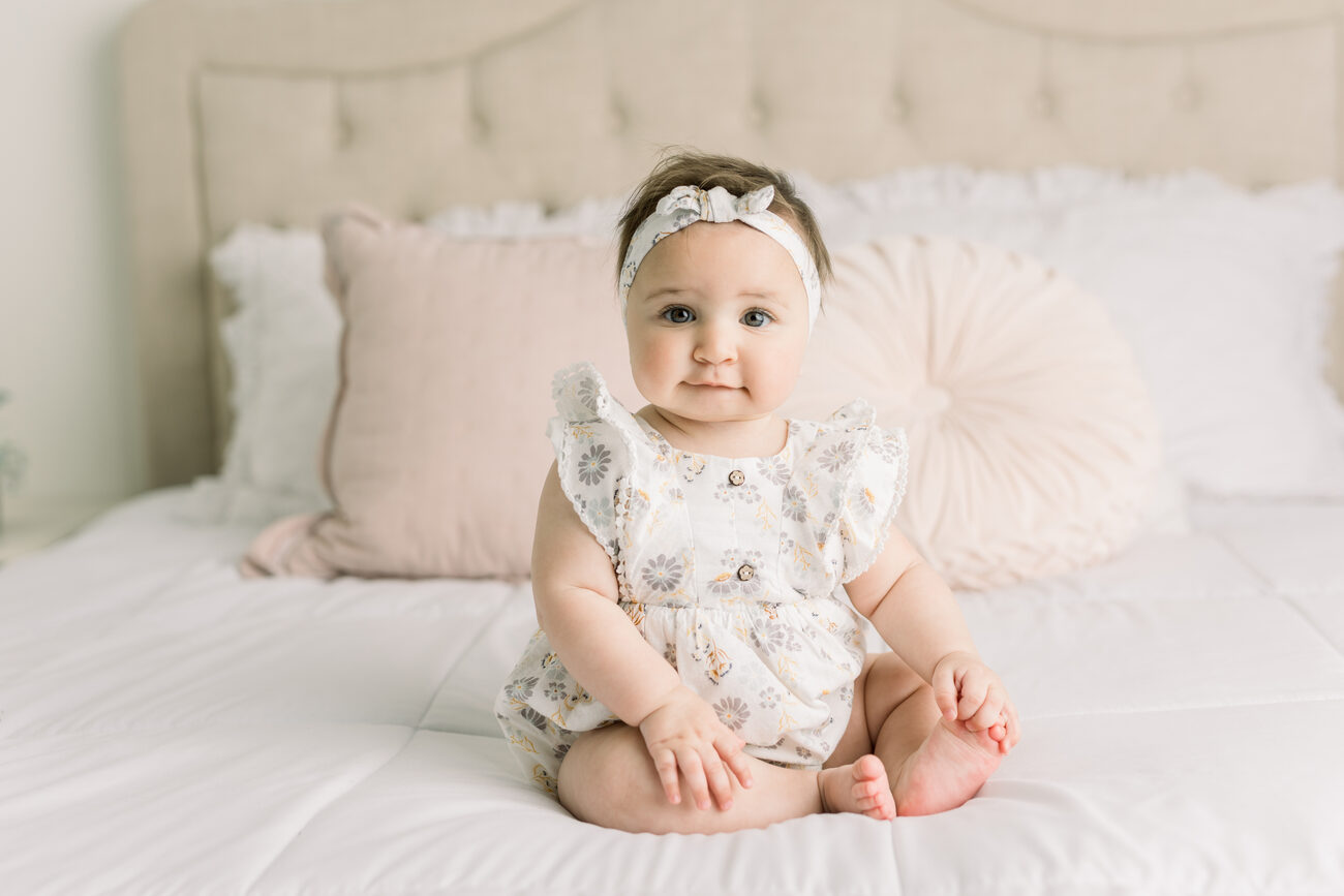 Baby girl in floral romper and headband sitting on bed, Boise sitter session photos