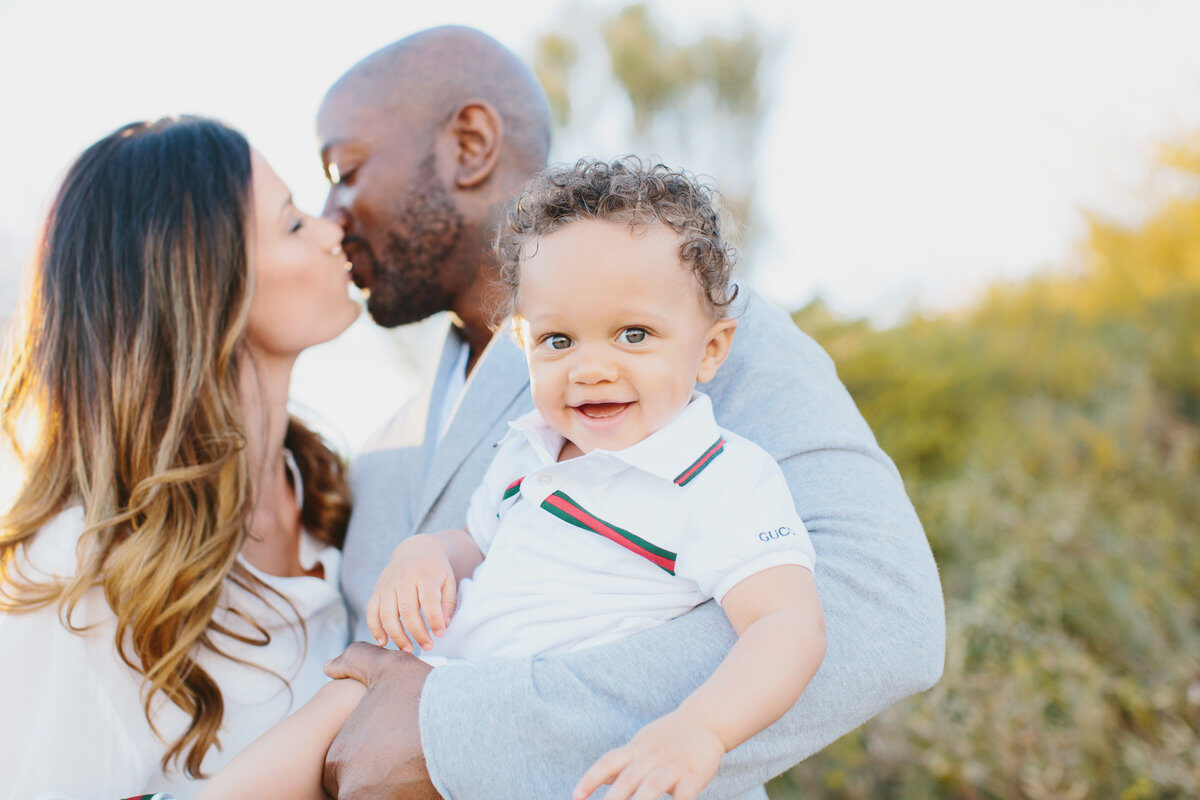 Mom and dad kissing while baby smiles at the camera, outdoor family session in Boise