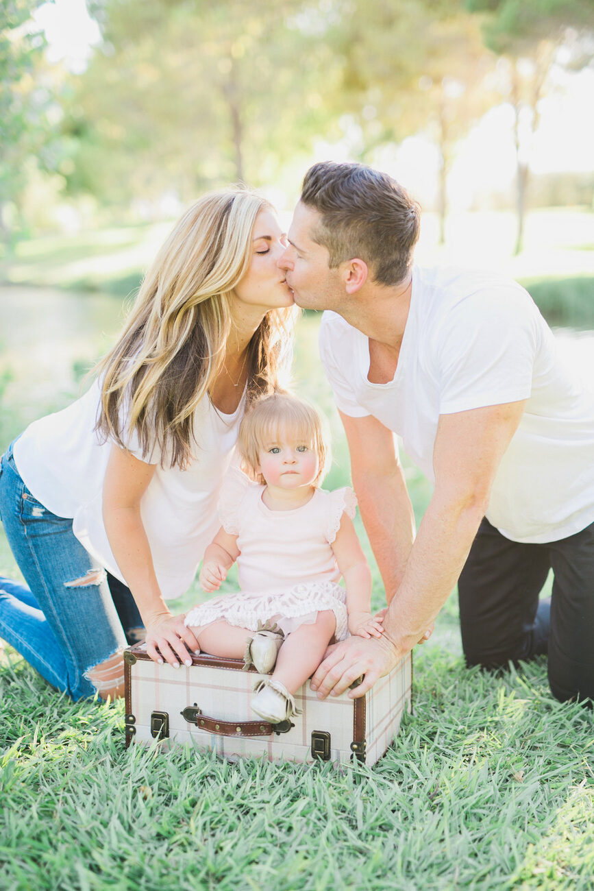 Parents kissing with toddler at vintage suitcase picnic, outdoor family session