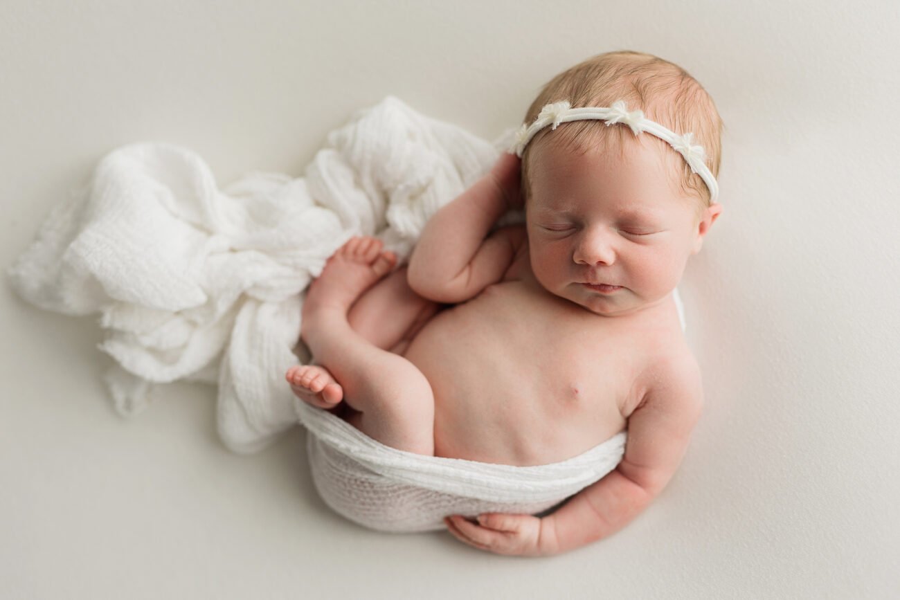Newborn baby girl in a floral headband curled up in a soft white wrap, Boise newborn session
