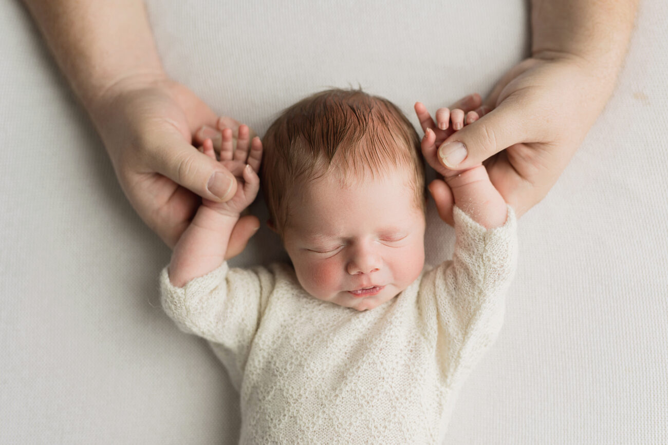 Parents holding their sleeping newborn’s hands overhead on a cream blanket, Boise newborn photographer